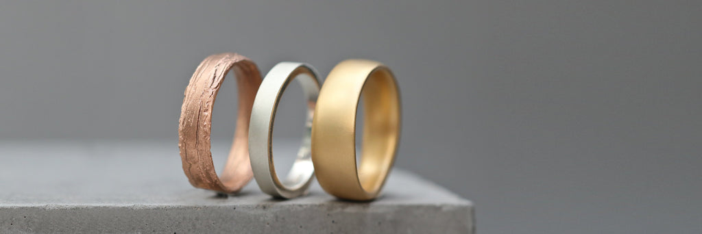 Mens wedding band rings shown in rose gold, white gold and yellow gold displayed on a concrete block.