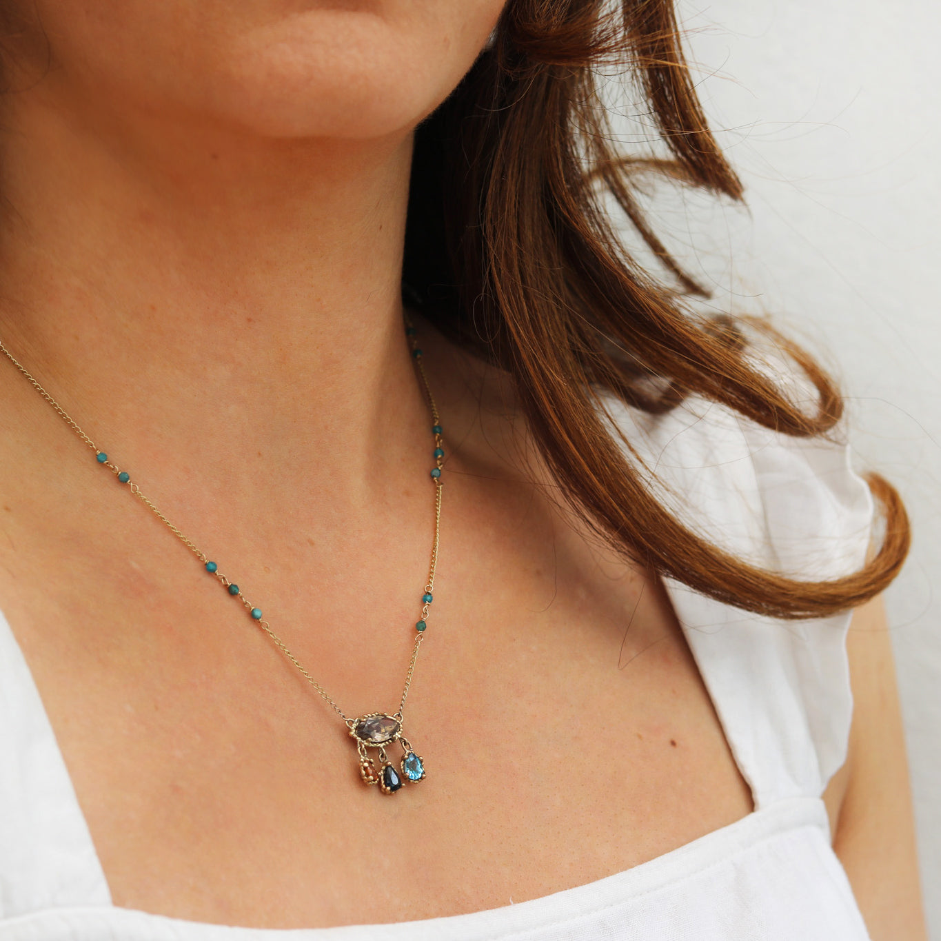 A close-up of a woman wearing a delicate gold chain necklace with small turquoise-blue beads and a matching multi-stone pendant.