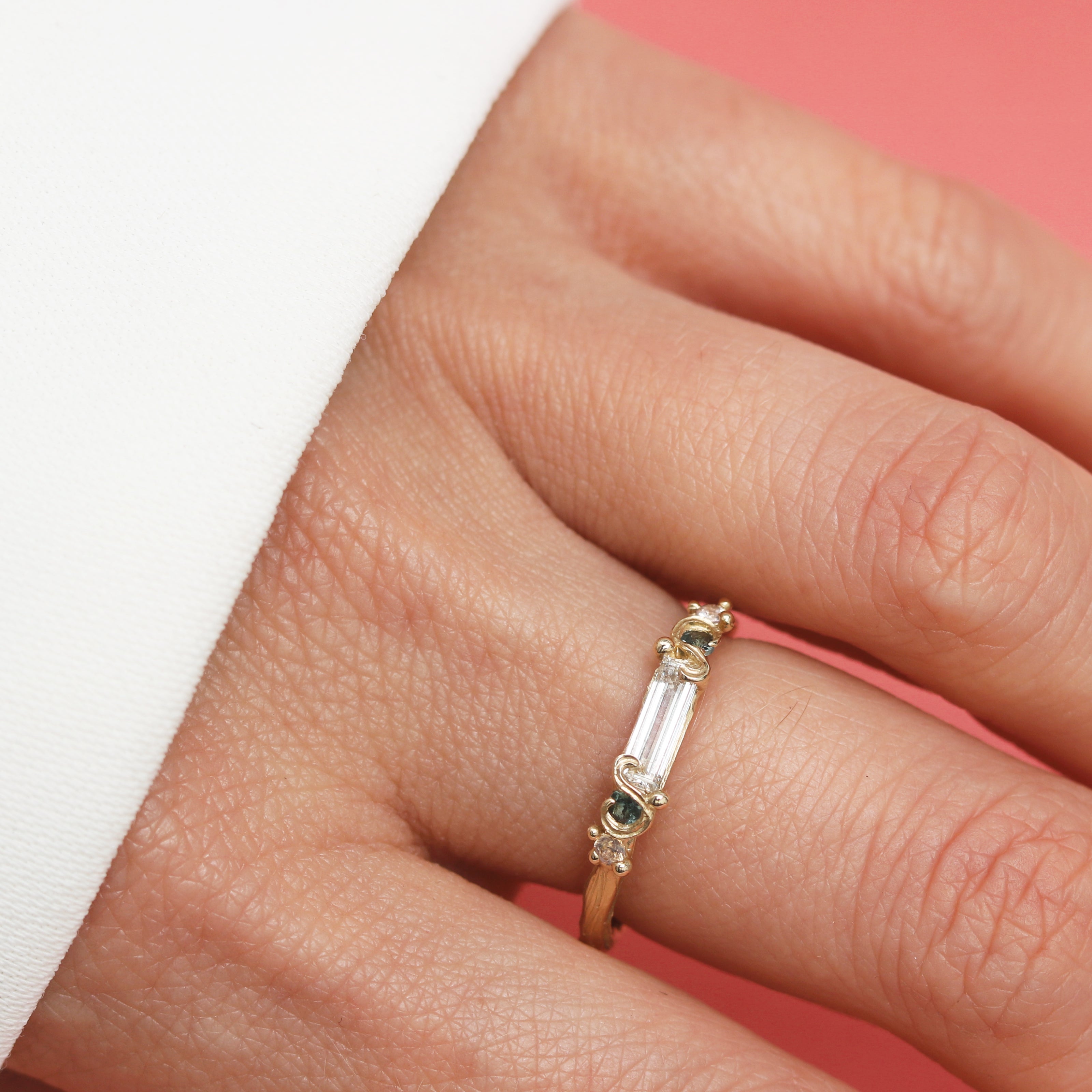 Close-up of a hand wearing a gold ring with a long white diamond and a green Sapphire on a pink background