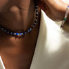 A close-up lifestyle shot of a model wearing a beaded choker necklace made of alternating dark blue and white beads, paired with a white top.