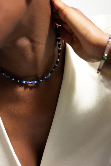 A close-up lifestyle shot of a model wearing a beaded choker necklace made of alternating dark blue and white beads, paired with a white top.
