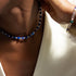A close-up lifestyle shot of a model wearing a beaded choker necklace made of alternating dark blue and white beads, paired with a white top.