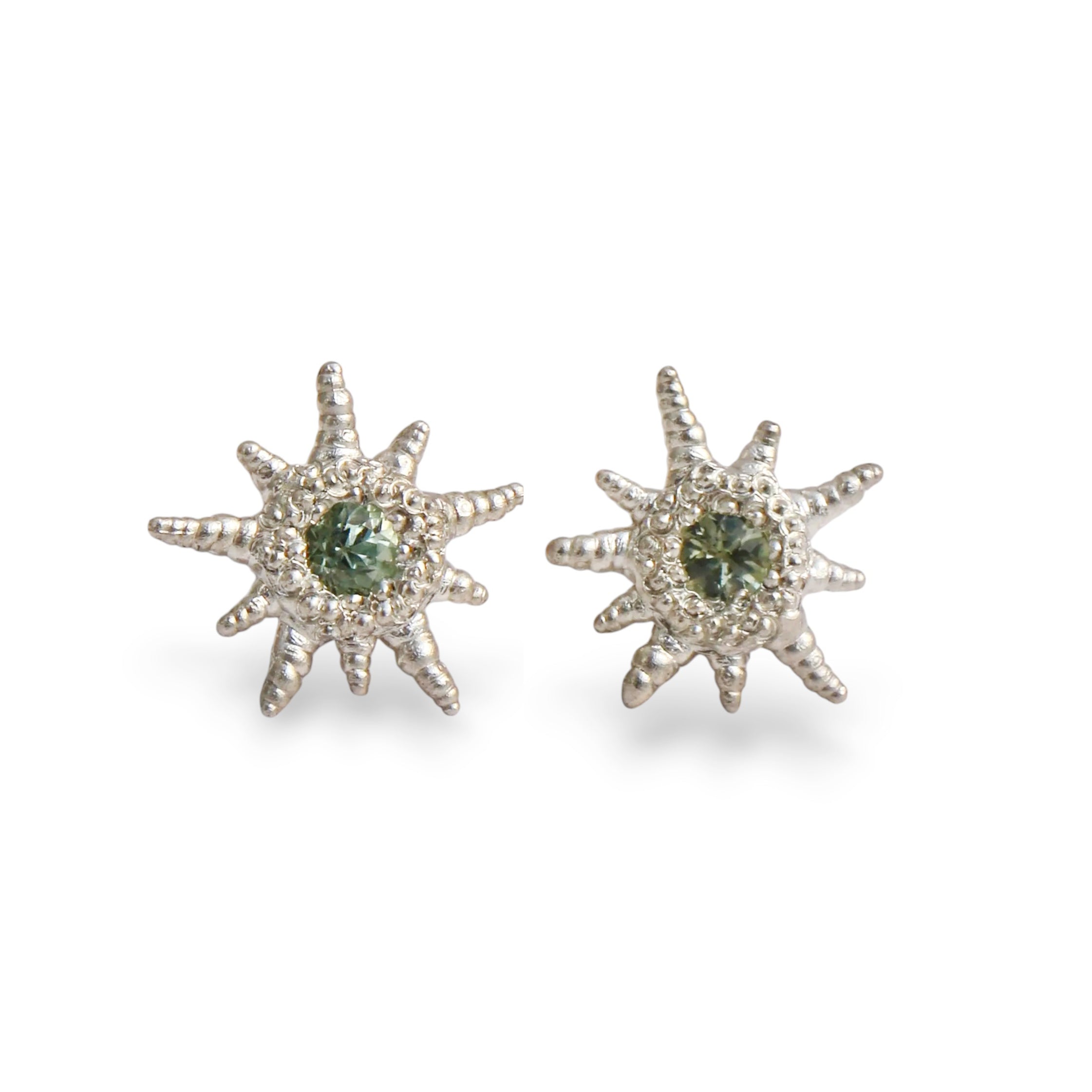 A pair of silver starburst stud earrings with a central green gemstone surrounded by a halo of small clear stones, set against a plain white background.