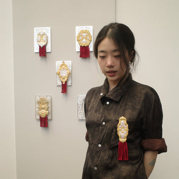 Woman wearing a dark brown dress with decorative brooches against a plain wall.