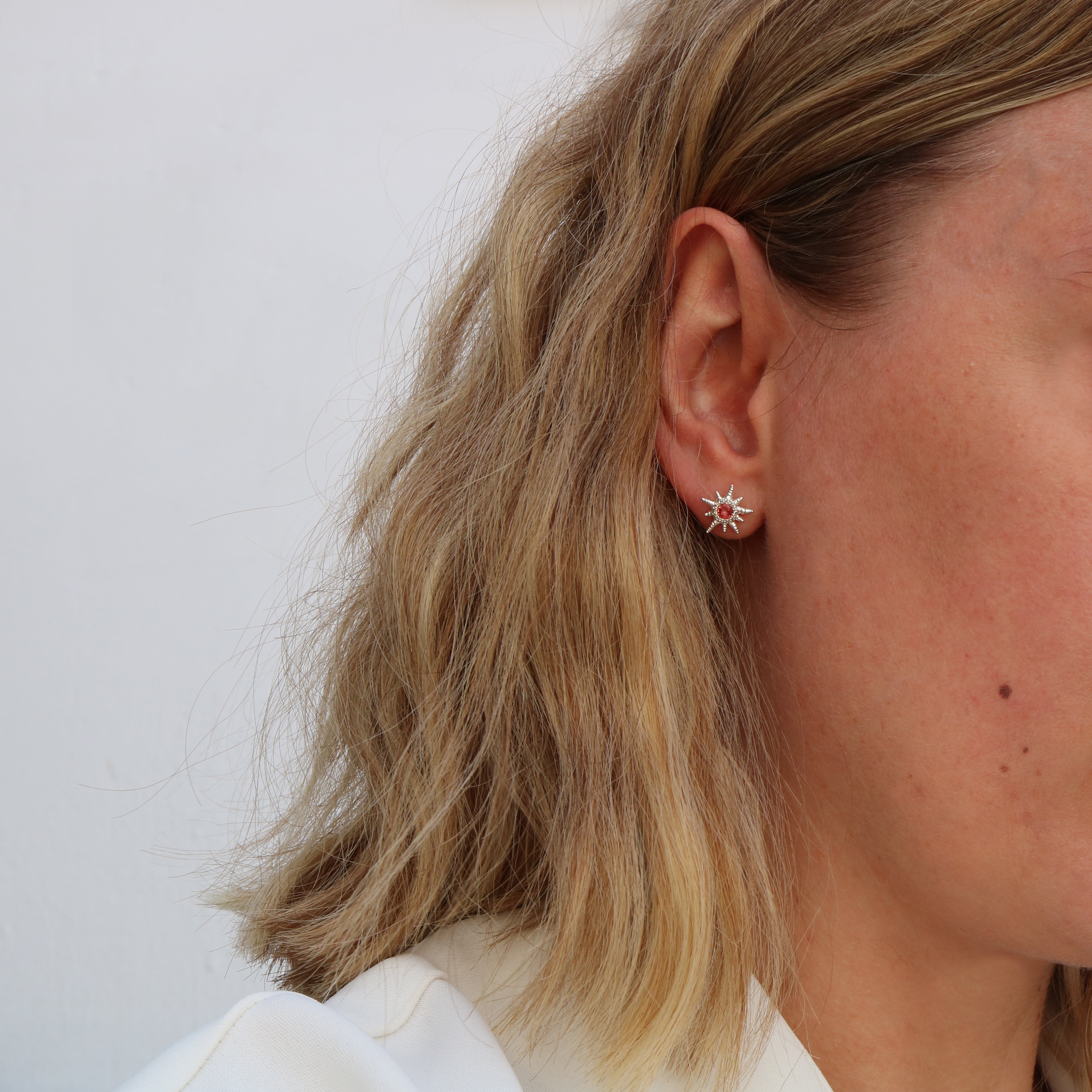 A close-up side profile of a person with blonde hair wearing a small silver-toned starburst stud earring against a plain light background.