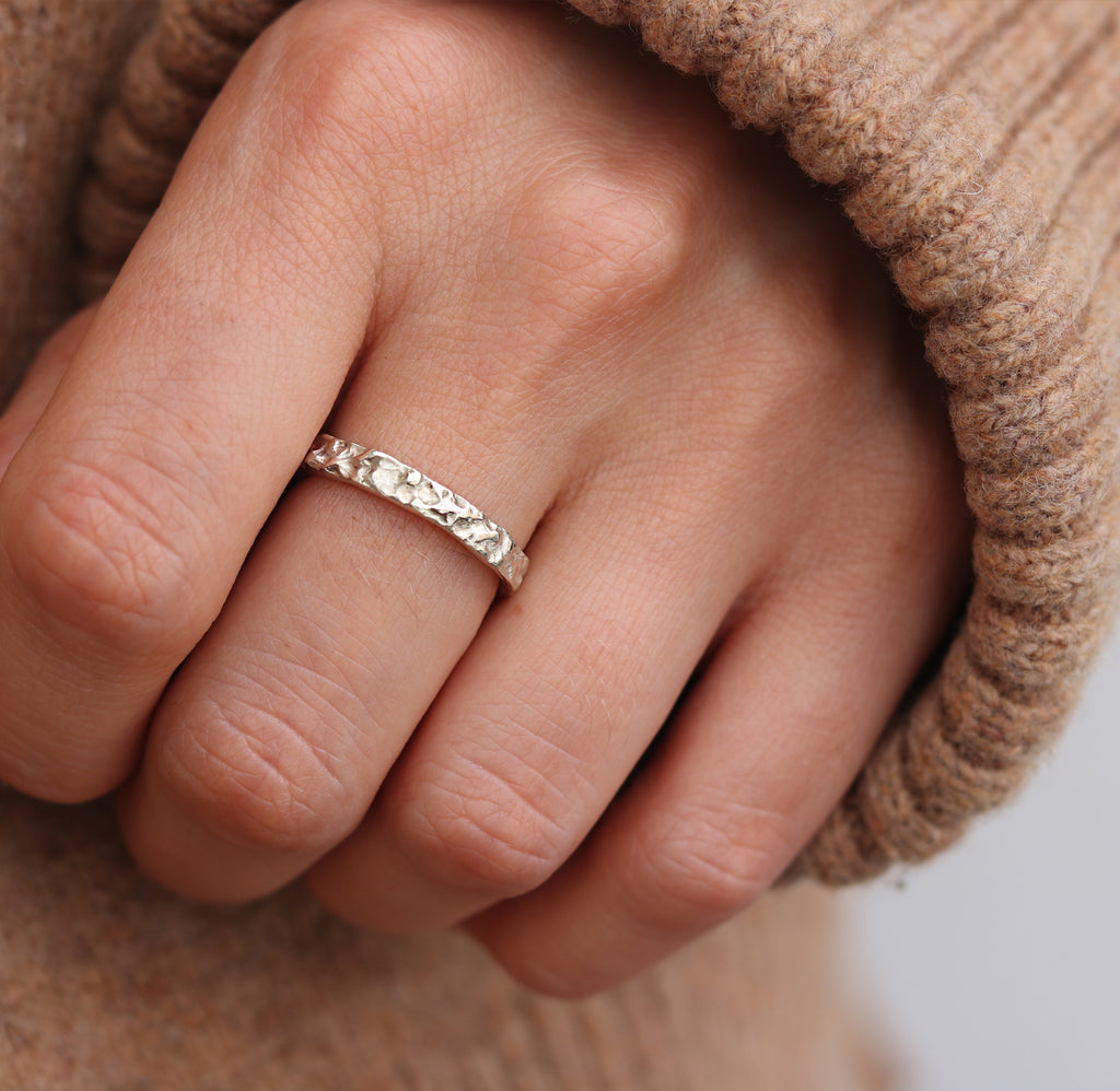 A narrow 9ct white gold wedding band with molten texture worn on womens hand with beige jumper