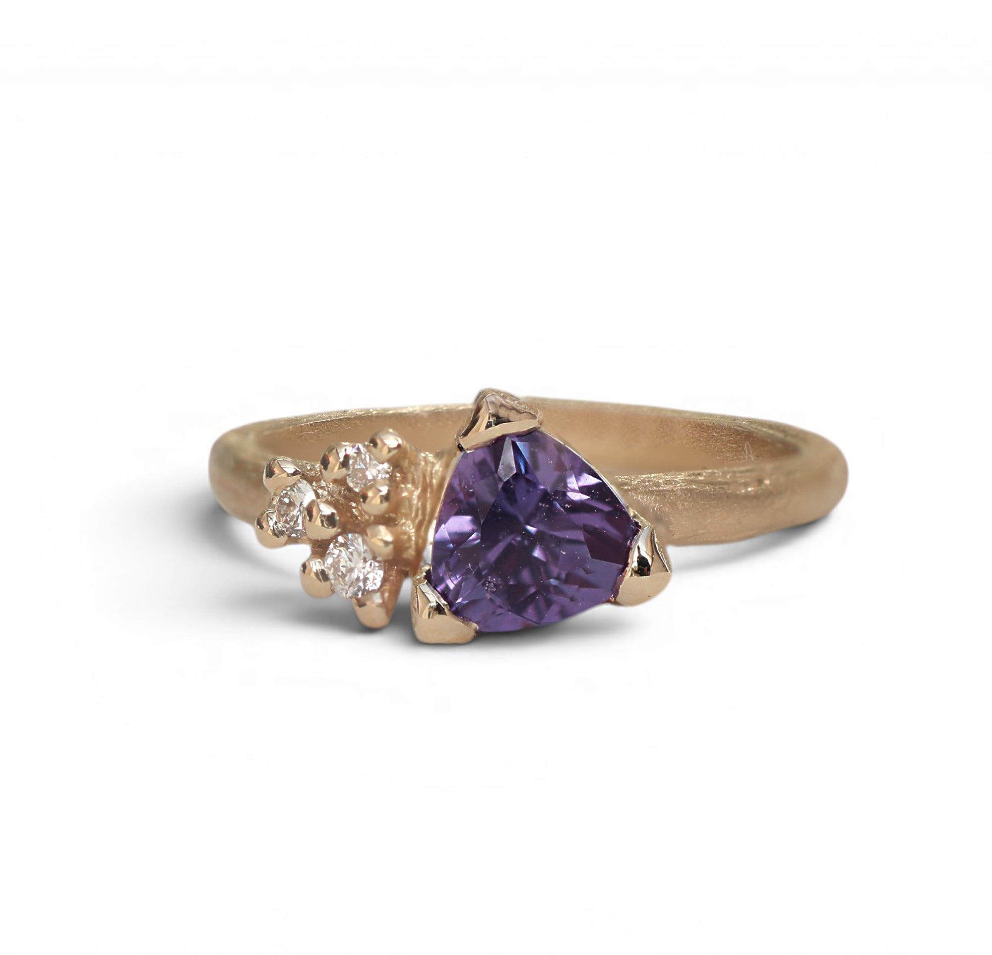 A gold ring featuring a large triangular purple gemstone in the center, accented by a cluster of small clear stones on one side, set against a clean white background.