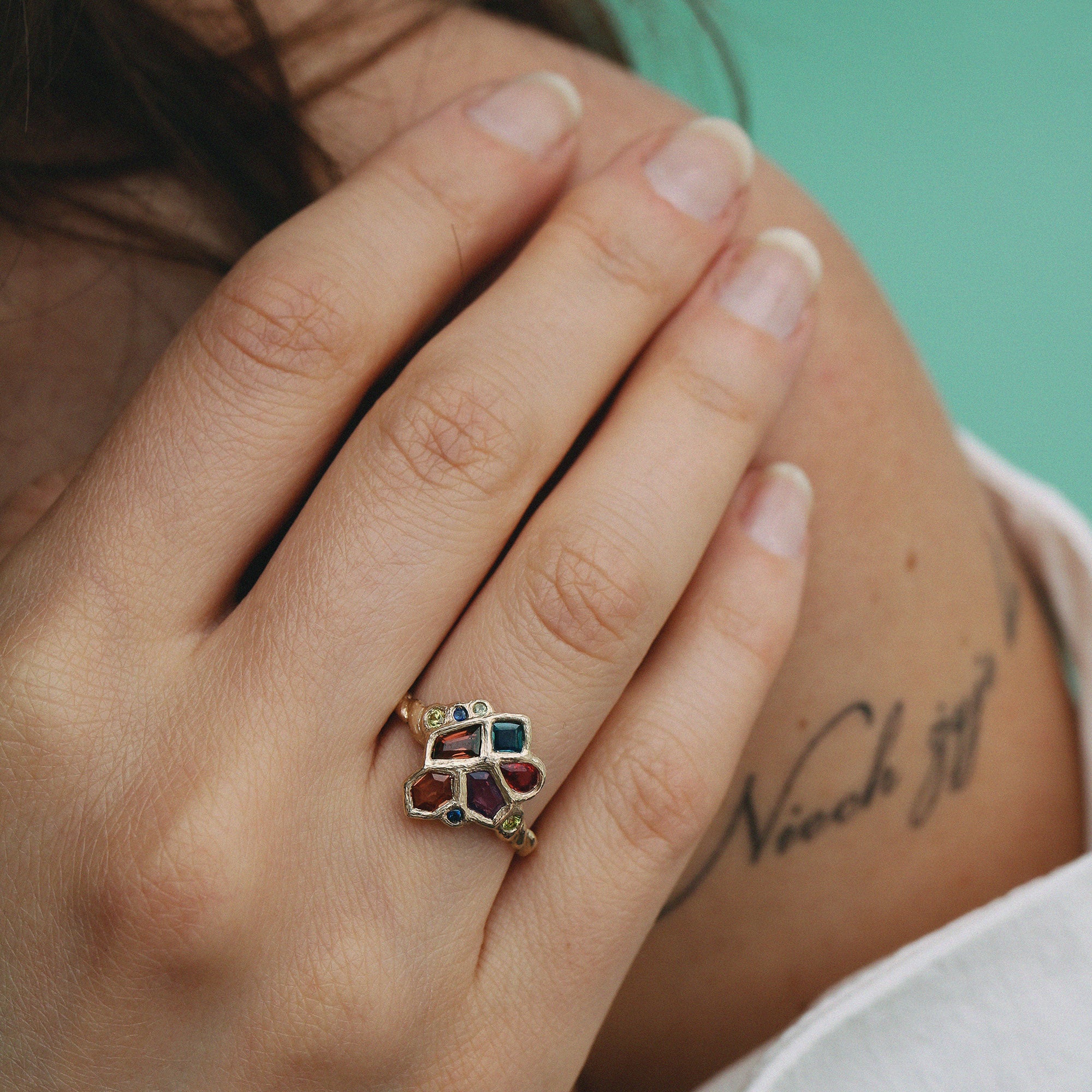 Close-up of a hand wearing a ring with colorful gemstones against a blurred background
