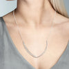 A close-up of a model wearing a delicate silver necklace with a curved, pavé-set pendant. The model has blonde hair and is wearing a gray V-neck top against a neutral gray background.