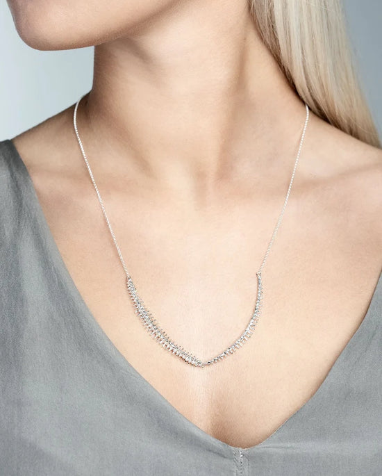 A close-up of a model wearing a delicate silver necklace with a curved, pavé-set pendant. The model has blonde hair and is wearing a gray V-neck top against a neutral gray background.