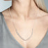 A close-up of a model wearing a delicate silver necklace with a curved, pavé-set pendant. The model has blonde hair and is wearing a gray V-neck top against a neutral gray background.