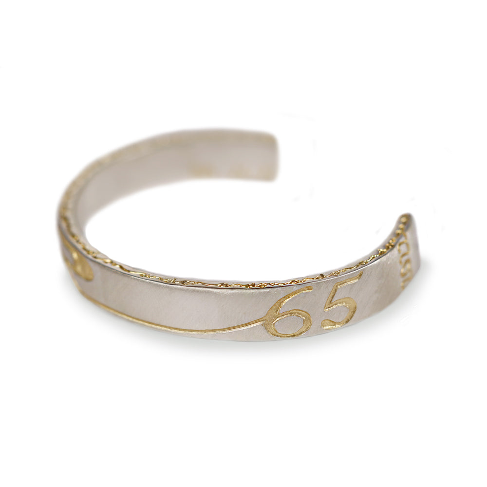 A silver-toned open cuff bracelet with gold trim and the number '65' engraved on the front, shown on a plain white background.