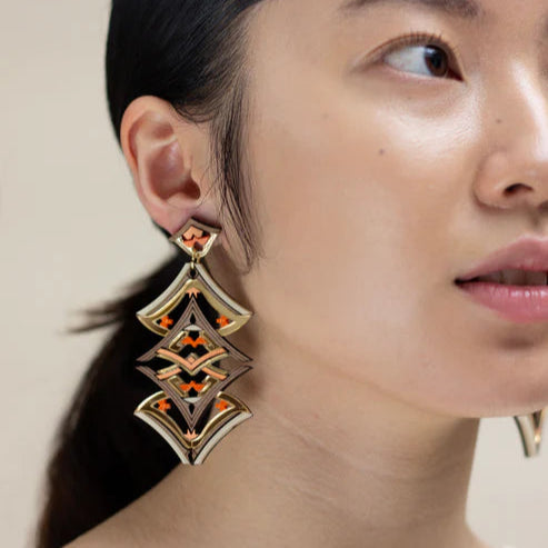 Close-up of a woman wearing gold and orange earrings against a neutral background
