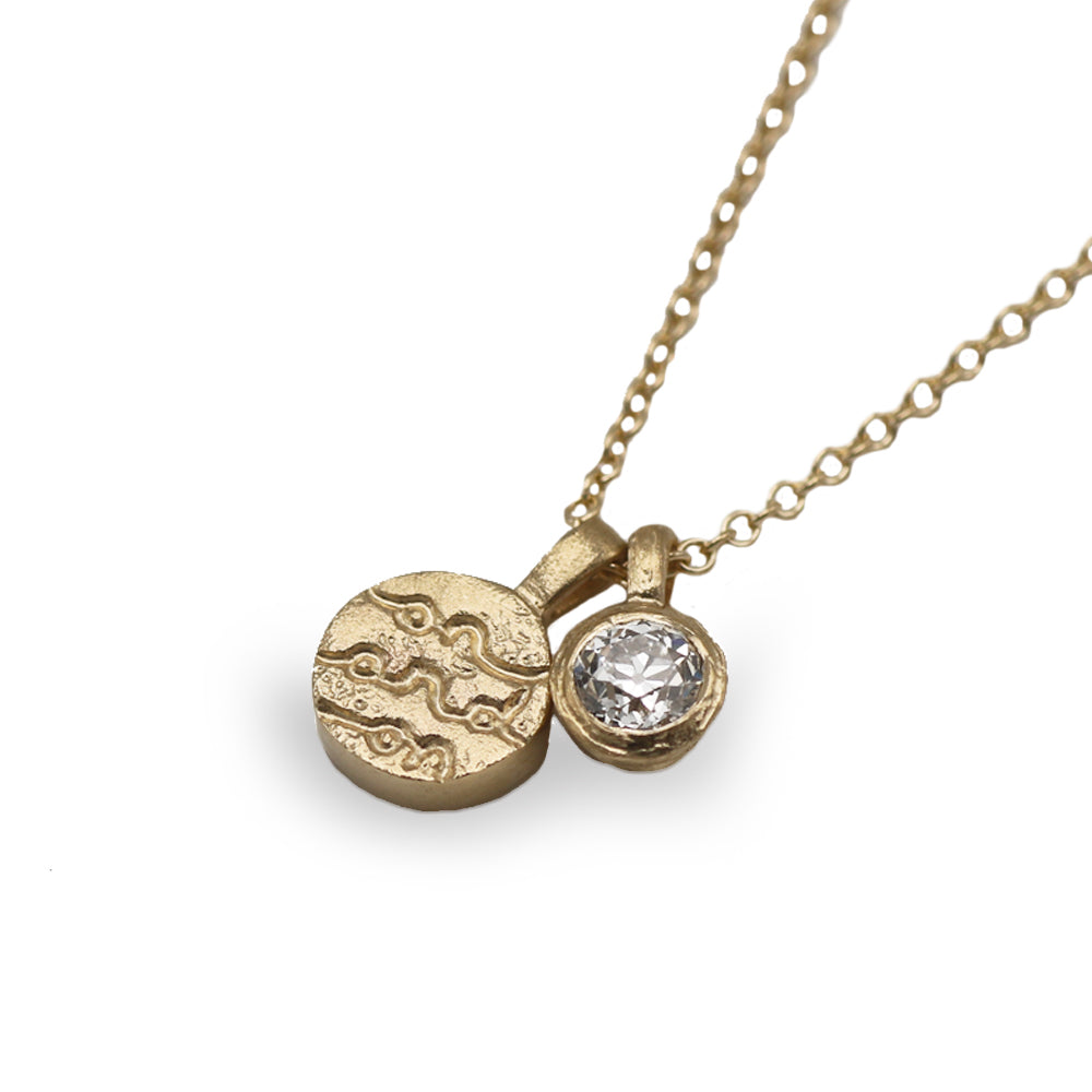 A gold pendant necklace featuring a round, textured gold disc charm and a smaller bezel-set clear gemstone accent, shown on a white background.