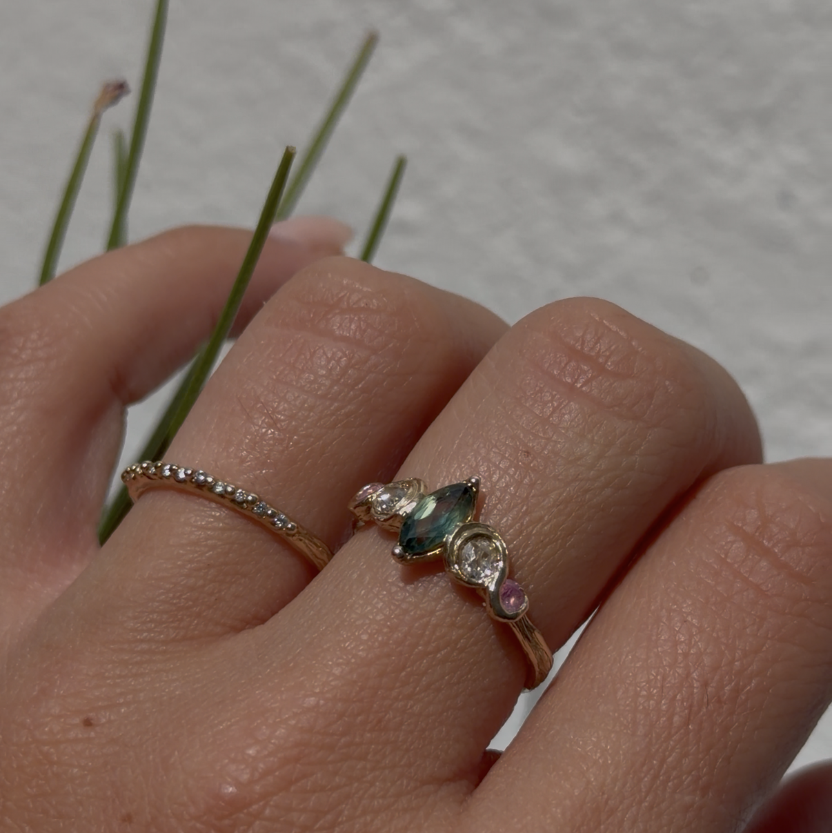 A close-up of a hand wearing a delicate gold ring with a small green and clear gemstone cluster, alongside a thin gold band.