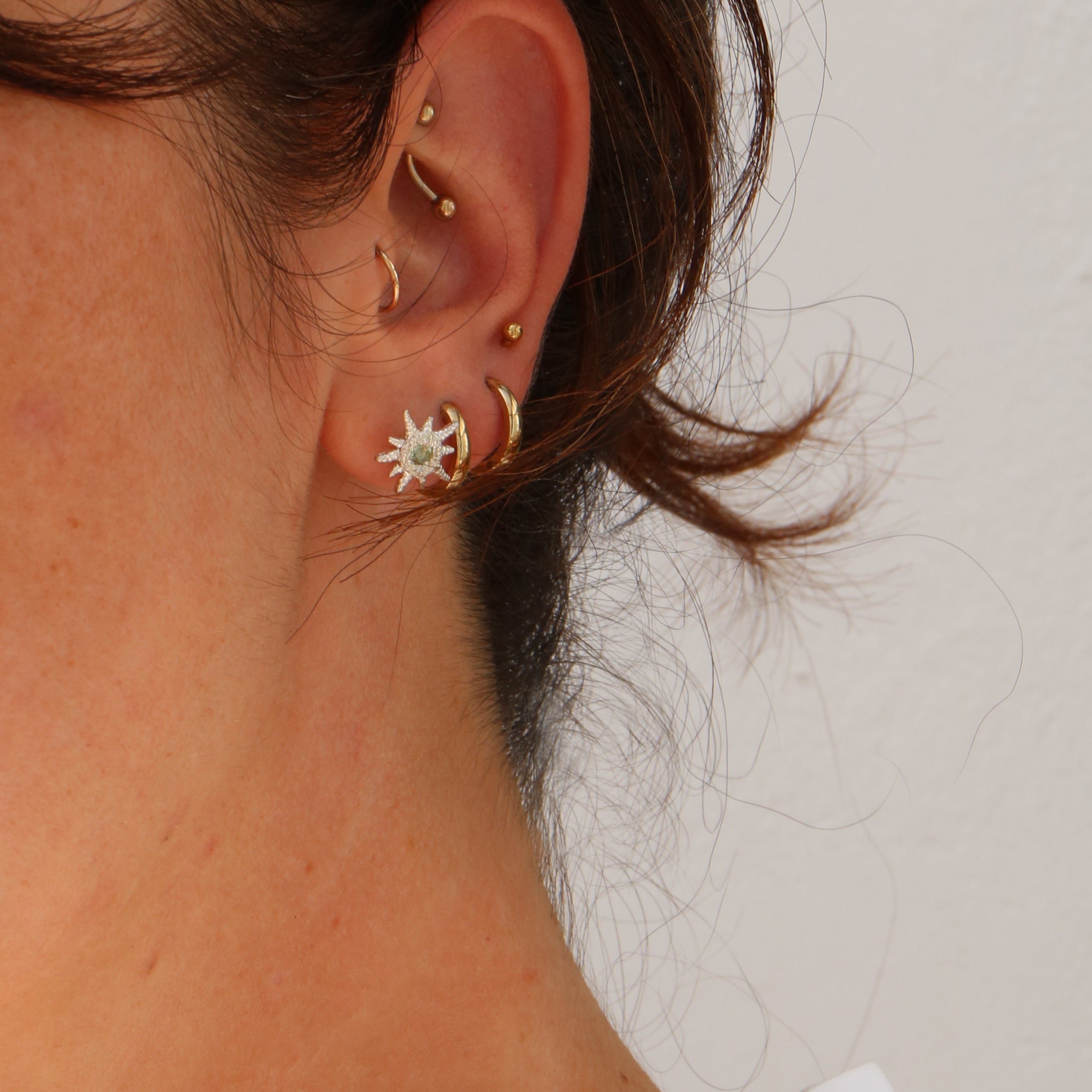 A close-up of a person's ear showcasing multiple gold ear piercings, including a starburst stud earring, a crescent moon hoop, and curved ear cuffs, against a plain off-white wall.