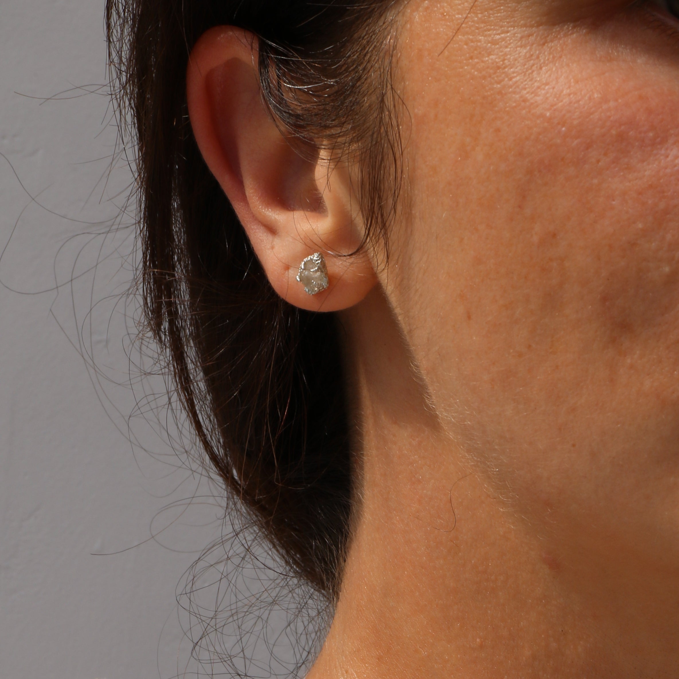 A close-up of a person's ear wearing a small, irregular-cut silver stud earring against a plain gray background.